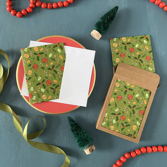 Christmas greeting cards patterned with midcentury-inspired ornaments, holly, and candy canes appear on a festive blue background with red beads, green ribbon, and bottle brush trees.