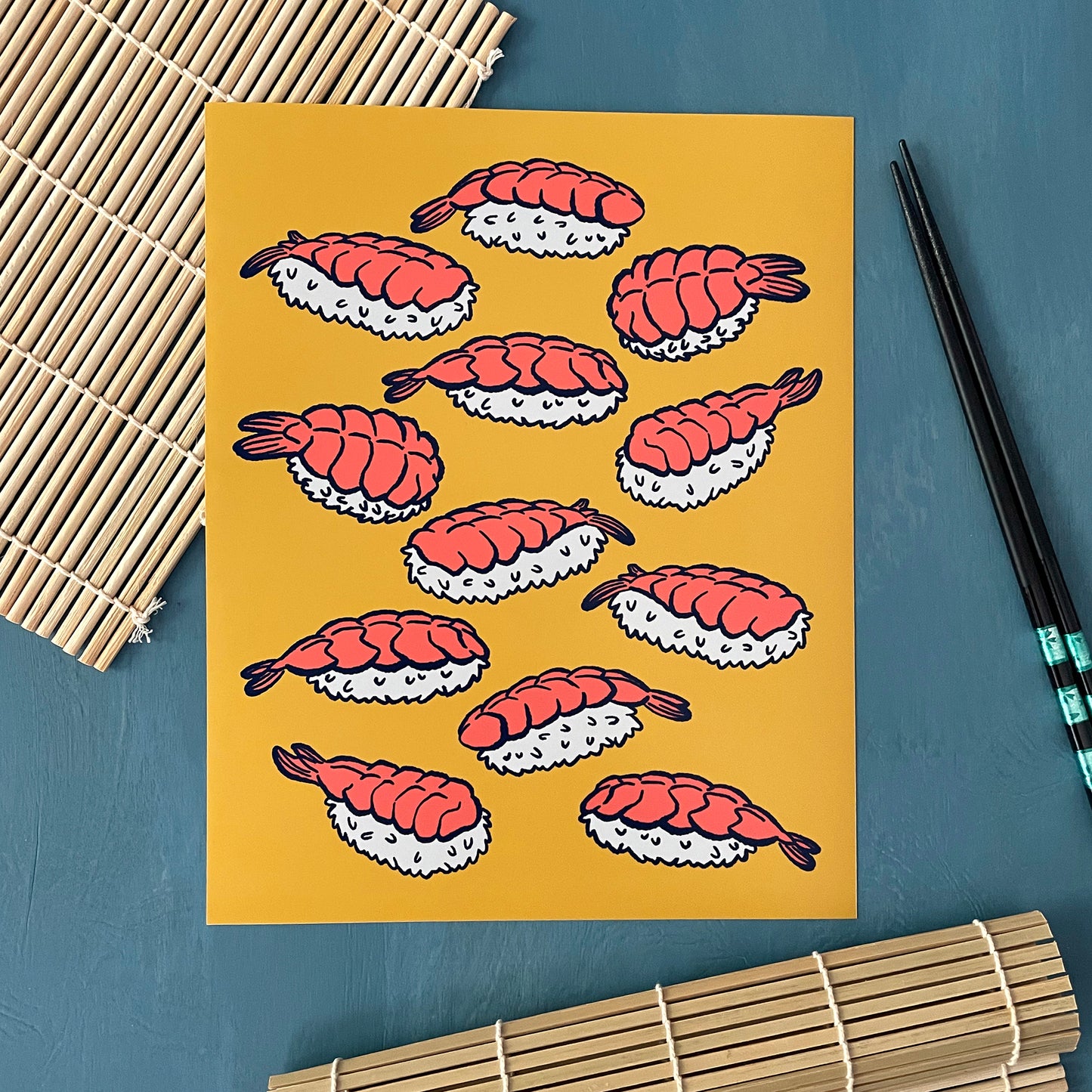 An art print shows bright pink shrimp on white rice outlined in navy blue against a yellow background. The art print is on a sushi mat and next to chopsticks.
