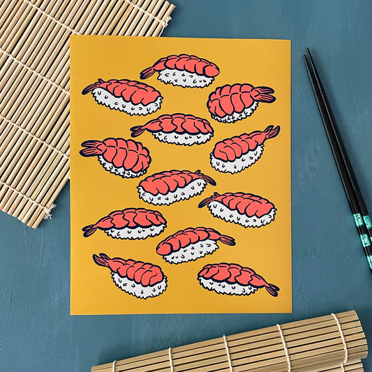 An art print shows bright pink shrimp on white rice outlined in navy blue against a yellow background. The art print is on a sushi mat and next to chopsticks.