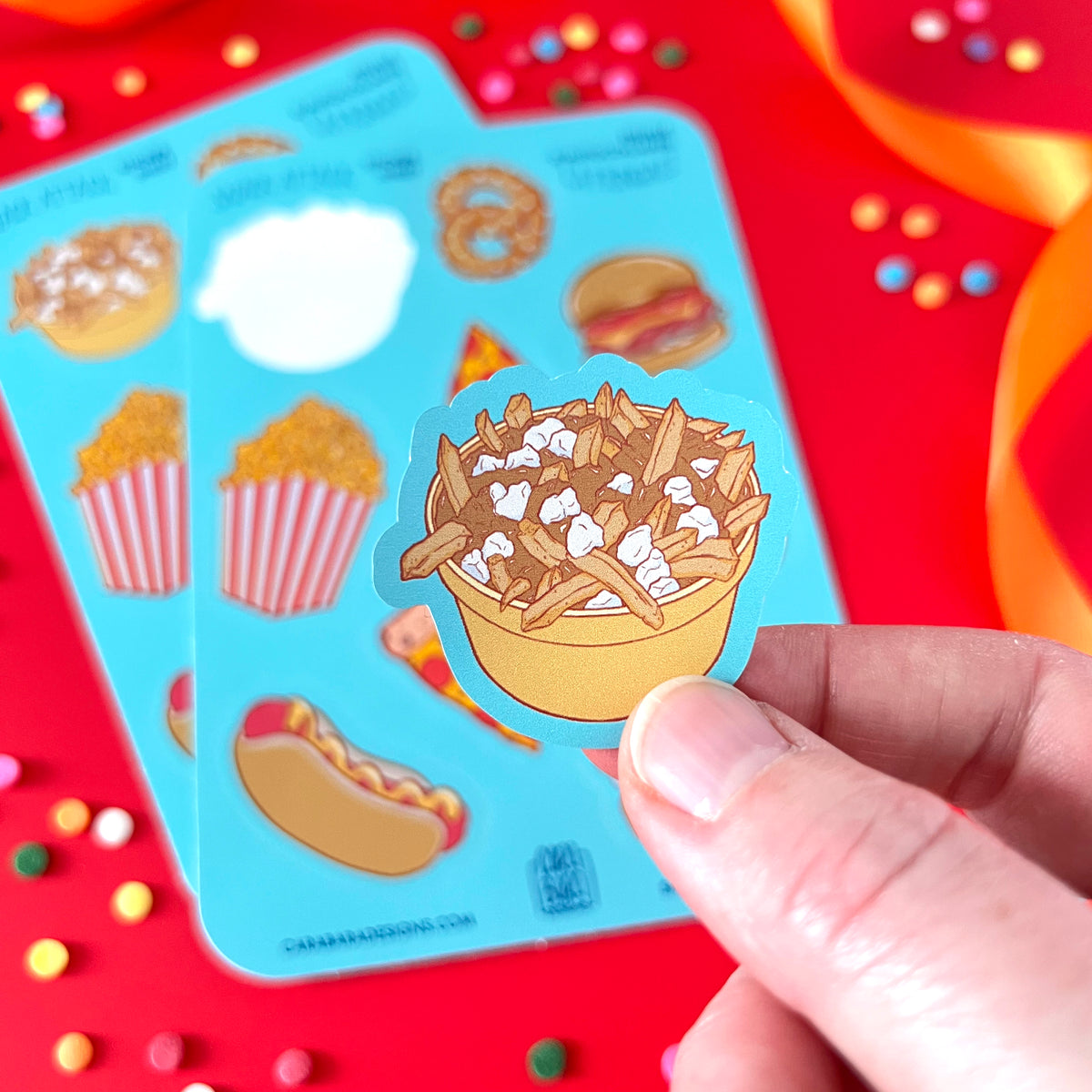 A hand holds a poutine sticker peeled from a sticker sheet. The food-themed sticker sheet is in the background.