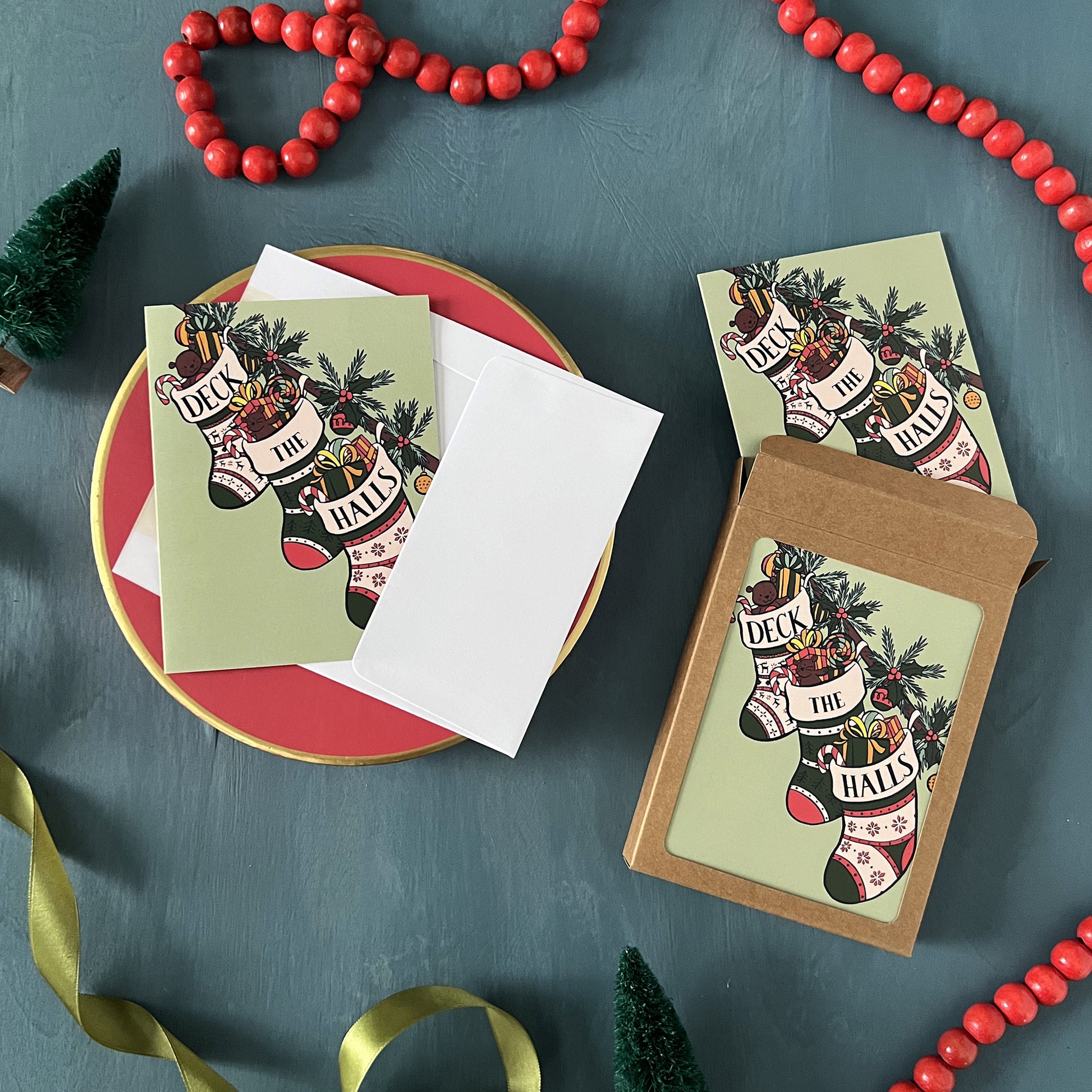 Christmas greeting cards with stockings reading Deck the Halls appear on a festive blue background with red beads, green ribbon, and bottle brush trees.