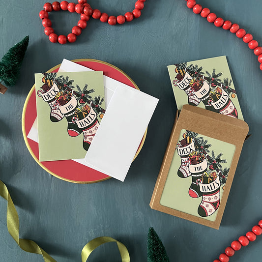 Christmas greeting cards with stockings reading Deck the Halls appear on a festive blue background with red beads, green ribbon, and bottle brush trees.