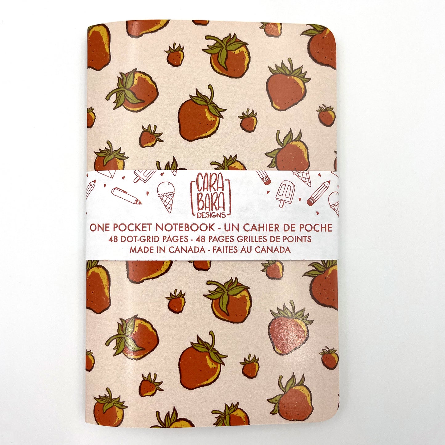 A small closed notebook has a blush pink cover that shows a scattered pattern of strawberries. The notebook is bound with a Carabara Designs belly band indicating in both French and English that it is a pocket notebook containing 48 dot grid pages and is made in Canada.