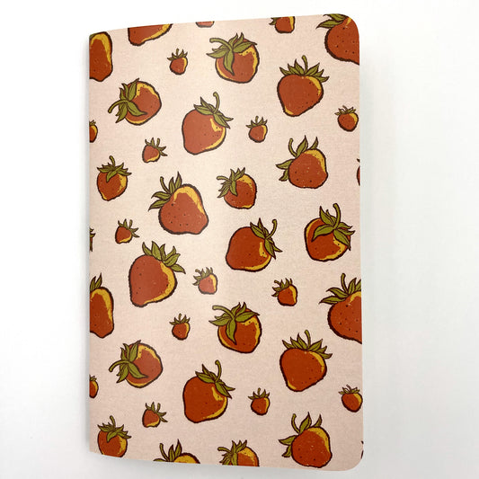 A small closed notebook has a blush pink cover that shows a scattered pattern of strawberries.