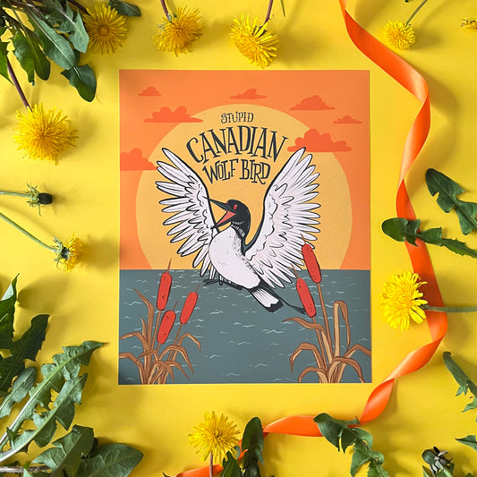 An art print shows a loon at sunset with cattails and the words 'stupid Canadian wolf bird' in hand lettering on a yellow background with ribbon and flowers.