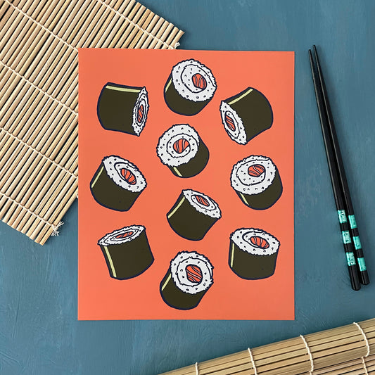 An art print depicts a cluster of seaweed-wrapped sushi rolls with salmon at the centre on an orange background. The print is on a sushi mat next to chopsticks.