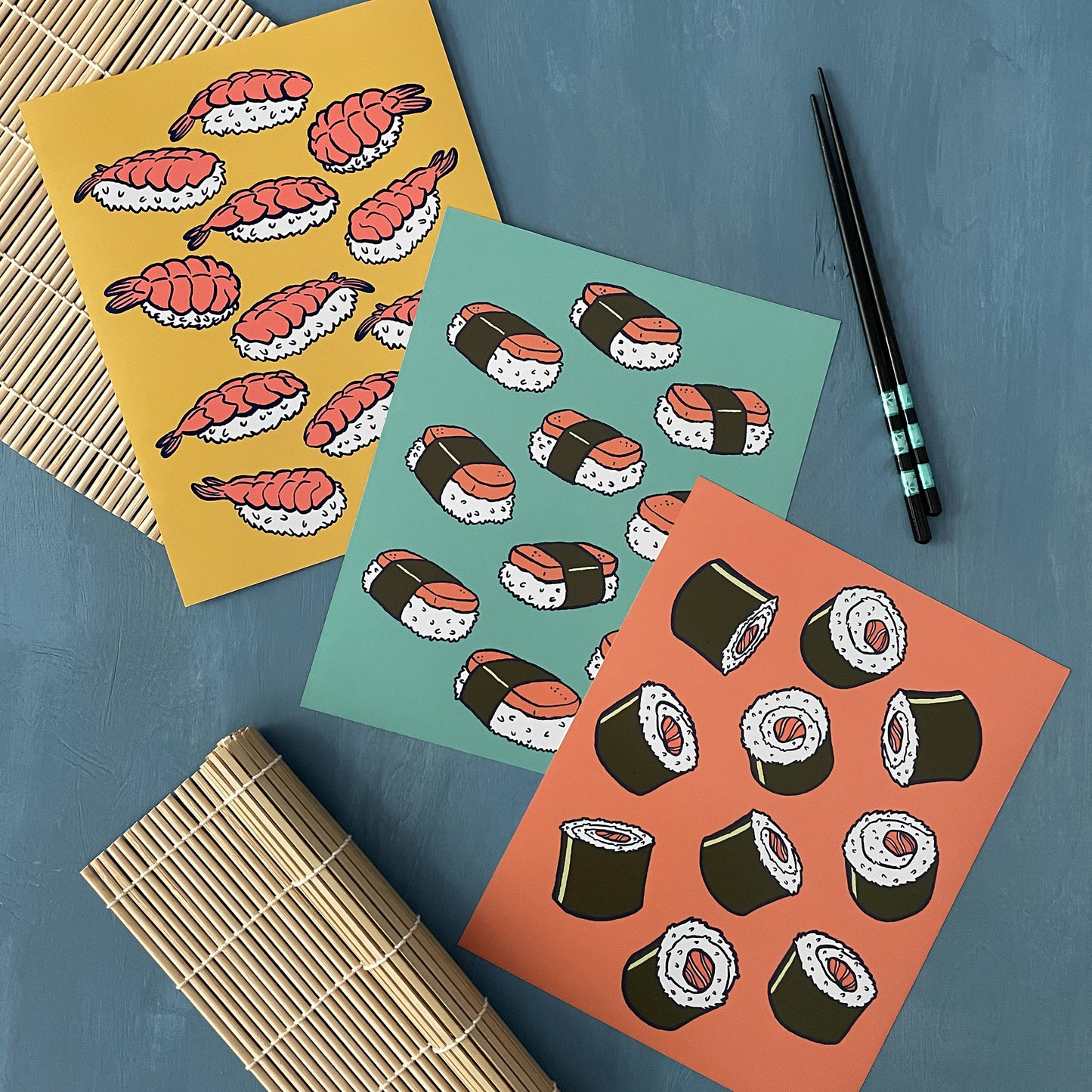 Three art prints, one illustrated with nigiri, one illustrated with musubi, and one illustrated with sushi rolls rest on a blue surface beside chopsticks and sushi mats.