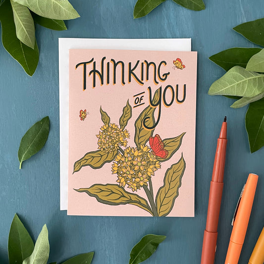 A pale pink greeting card has the words Thinking of You in dark green with a burst of milkweed blossoms and monarch butterflies below the letters. The card is surrounded by young milkweed leaves and brown, orange and yellow pens.