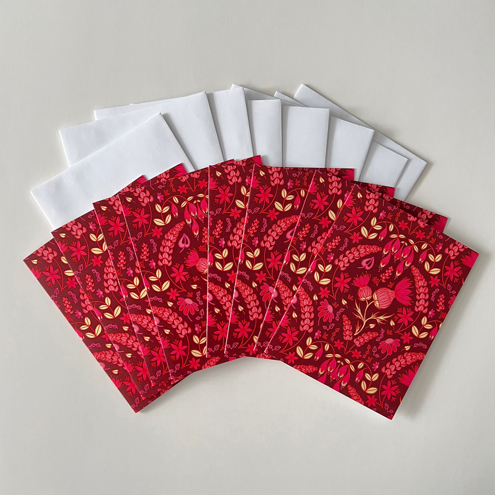 Eight burgundy greeting cards show a folk-style pattern of pink thistles and other wildflowers. The cards are on white envelopes.