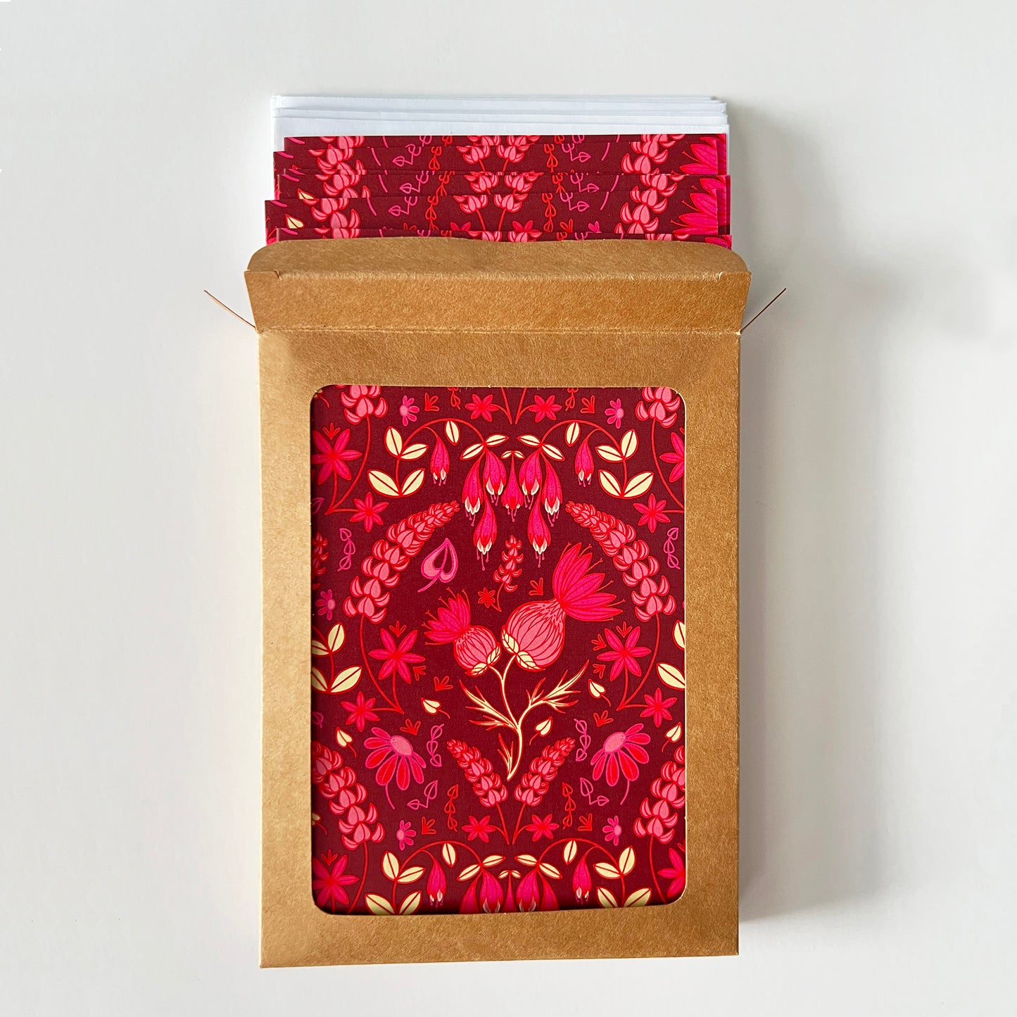 A kraft cardboard box with a window is open to show burgundy greeting cards with a folk-style pattern of pink thistles and other wildflowers. White envelopes peek out of the box.