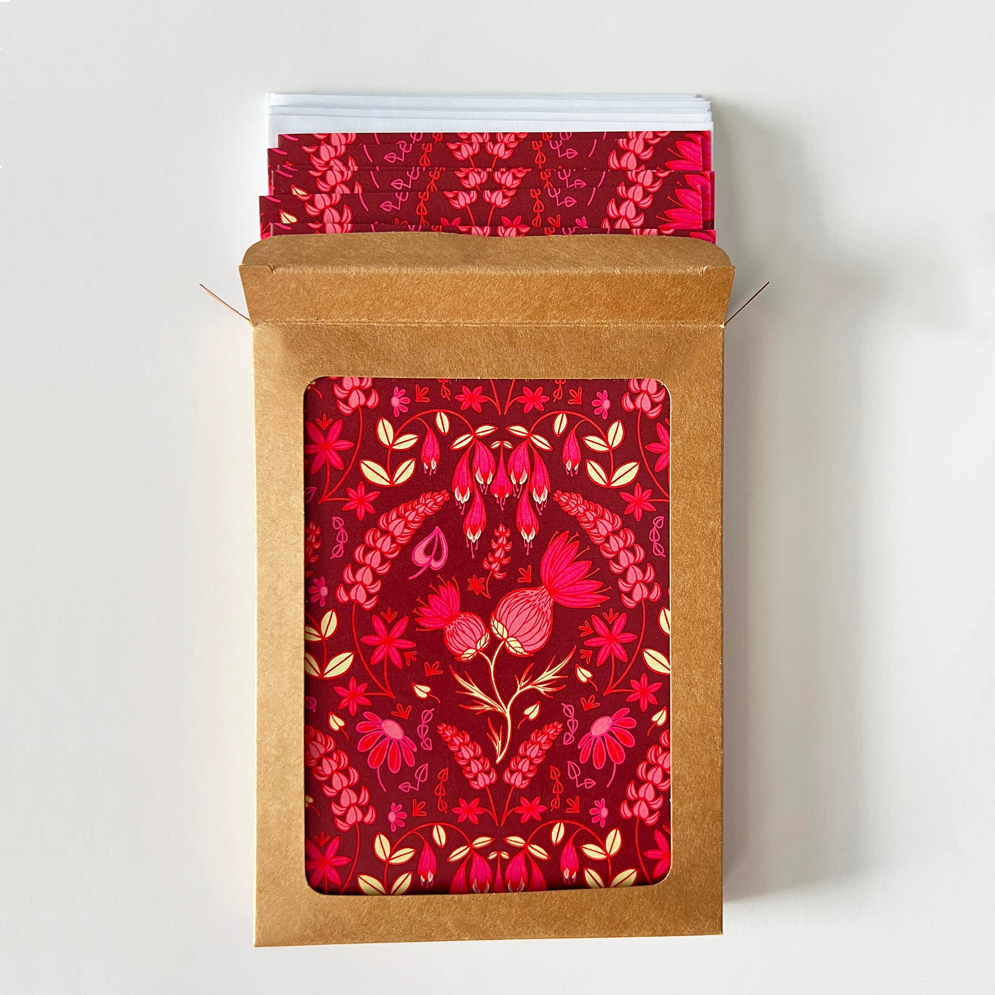 A kraft cardboard box with a window is open to show burgundy greeting cards with a folk-style pattern of pink thistles and other wildflowers. White envelopes peek out of the box.