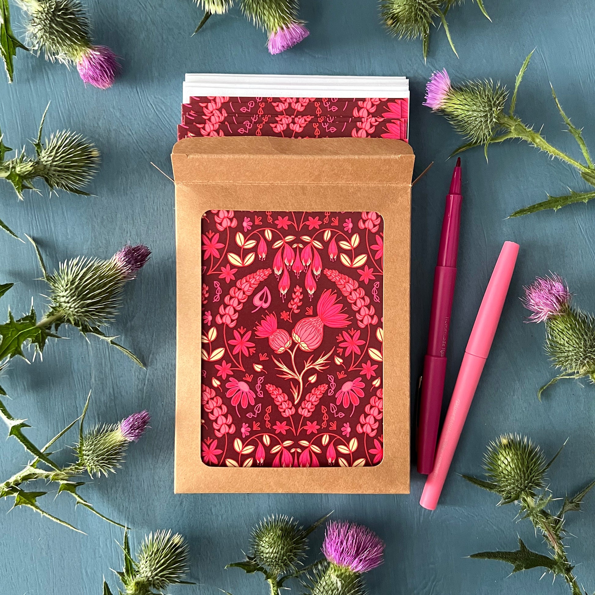 A kraft cardboard box with a window is open to show burgundy greeting cards with a folk-style pattern of pink thistles and other wildflowers. White envelopes peek out of the box which is surrounded by thistles.