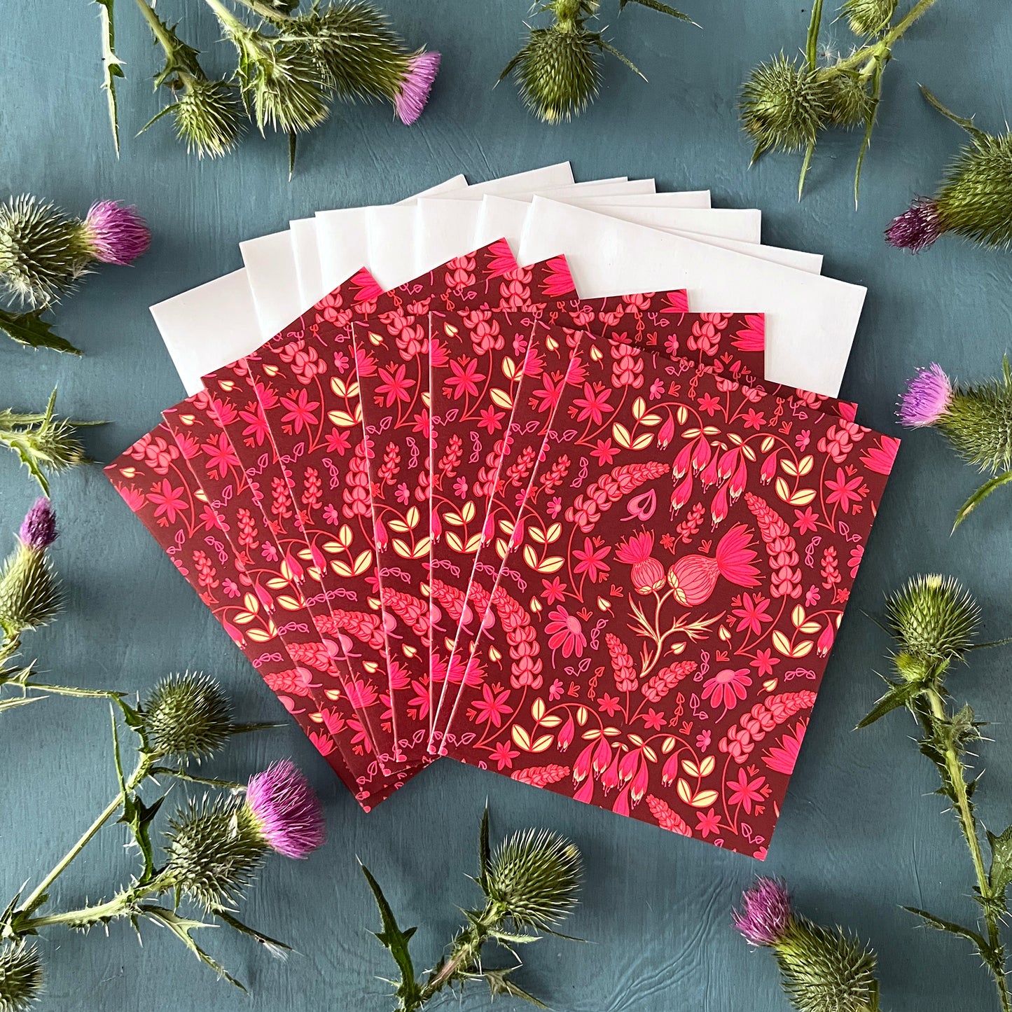 Eight burgundy greeting cards show a folk-style pattern of pink thistles and other wildflowers. The cards are on white envelopes and surrounded by thistles.