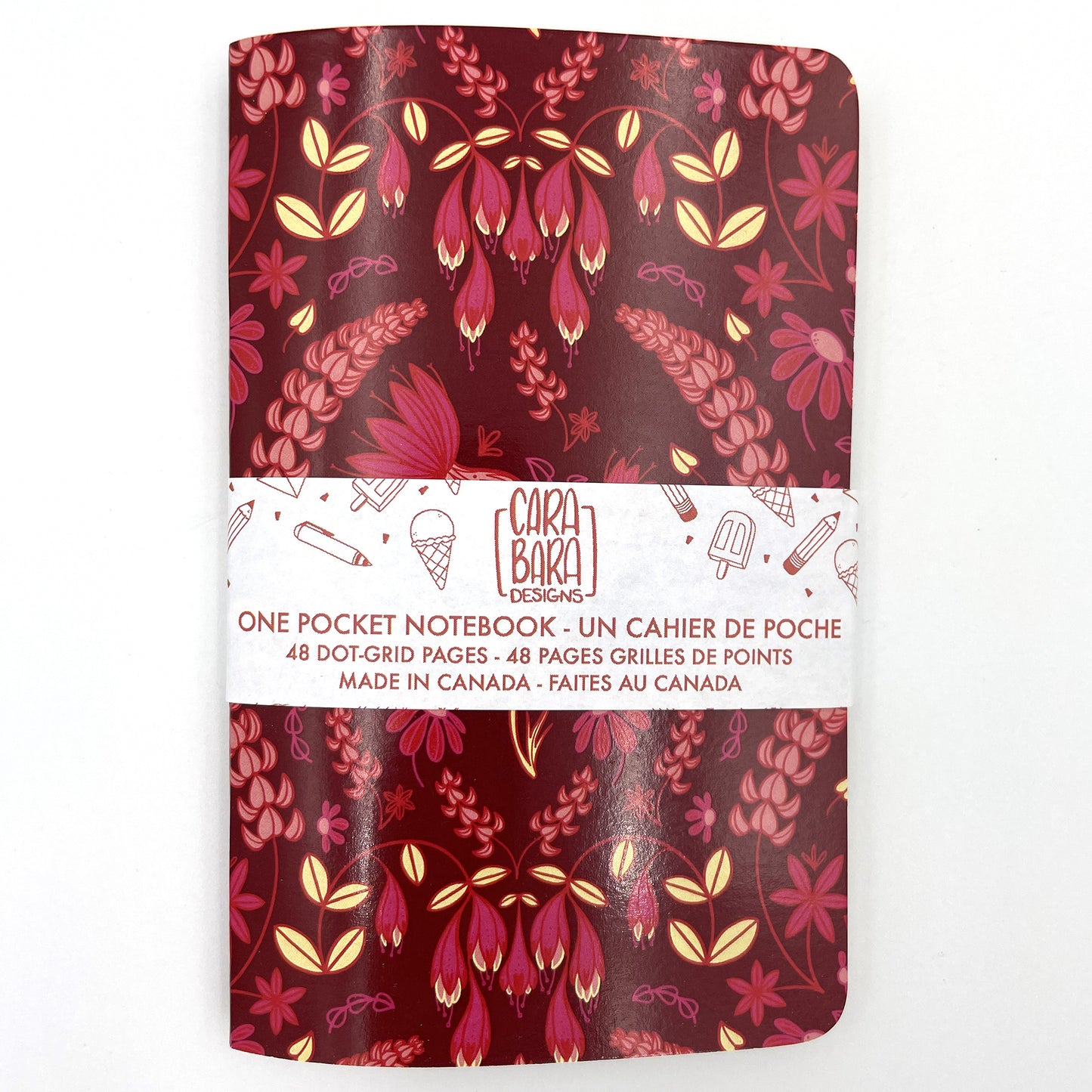 A small closed notebook has a burgundy cover that shows a pattern of thistles, echinacea, lupines and haskap blossoms. The notebook is bound with a Carabara Designs belly band indicating in both French and English that it is a pocket notebook containing 48 dot grid pages and is made in Canada.