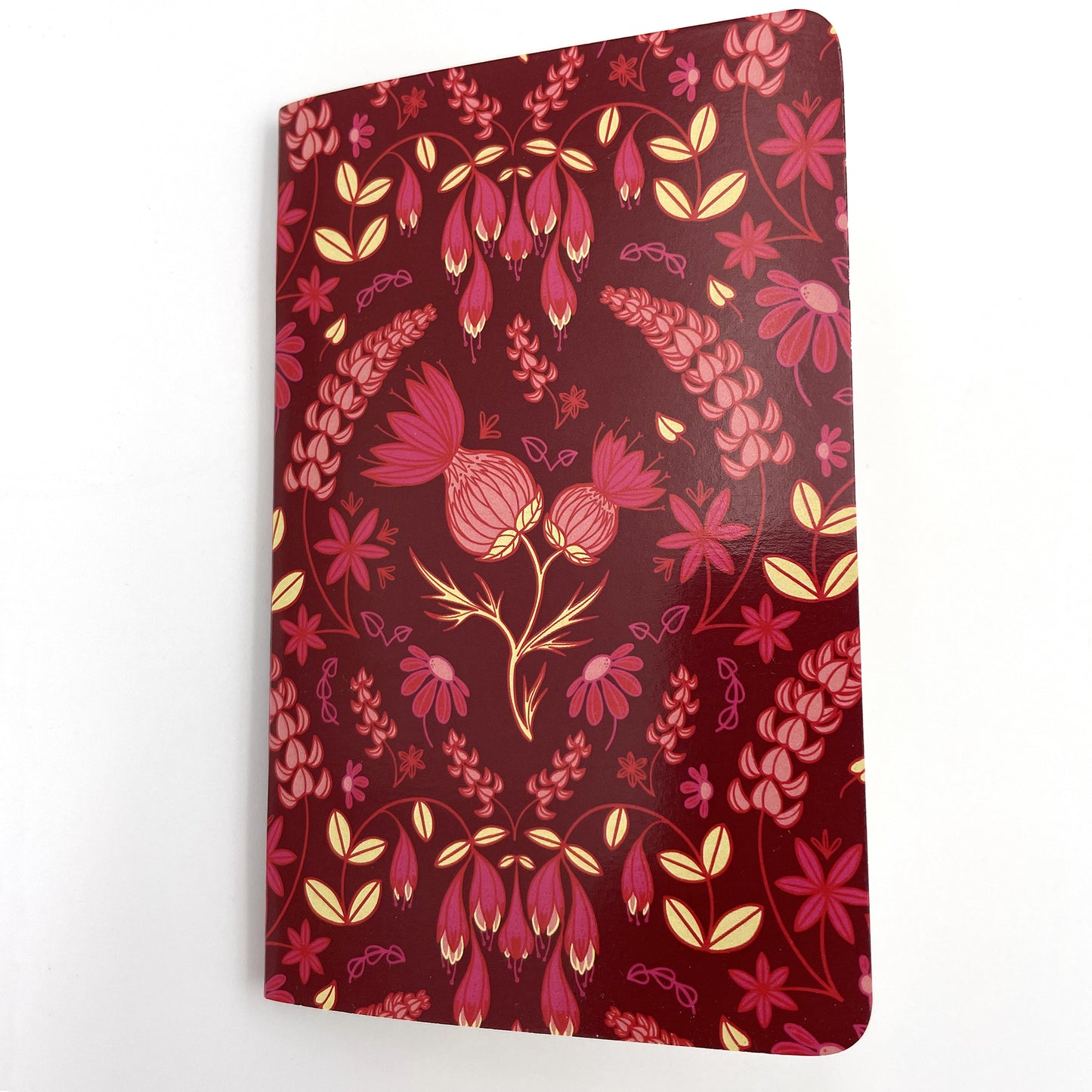 A small closed notebook has a burgundy cover that shows a pattern of thistles, echinacea, lupines and haskap blossoms.