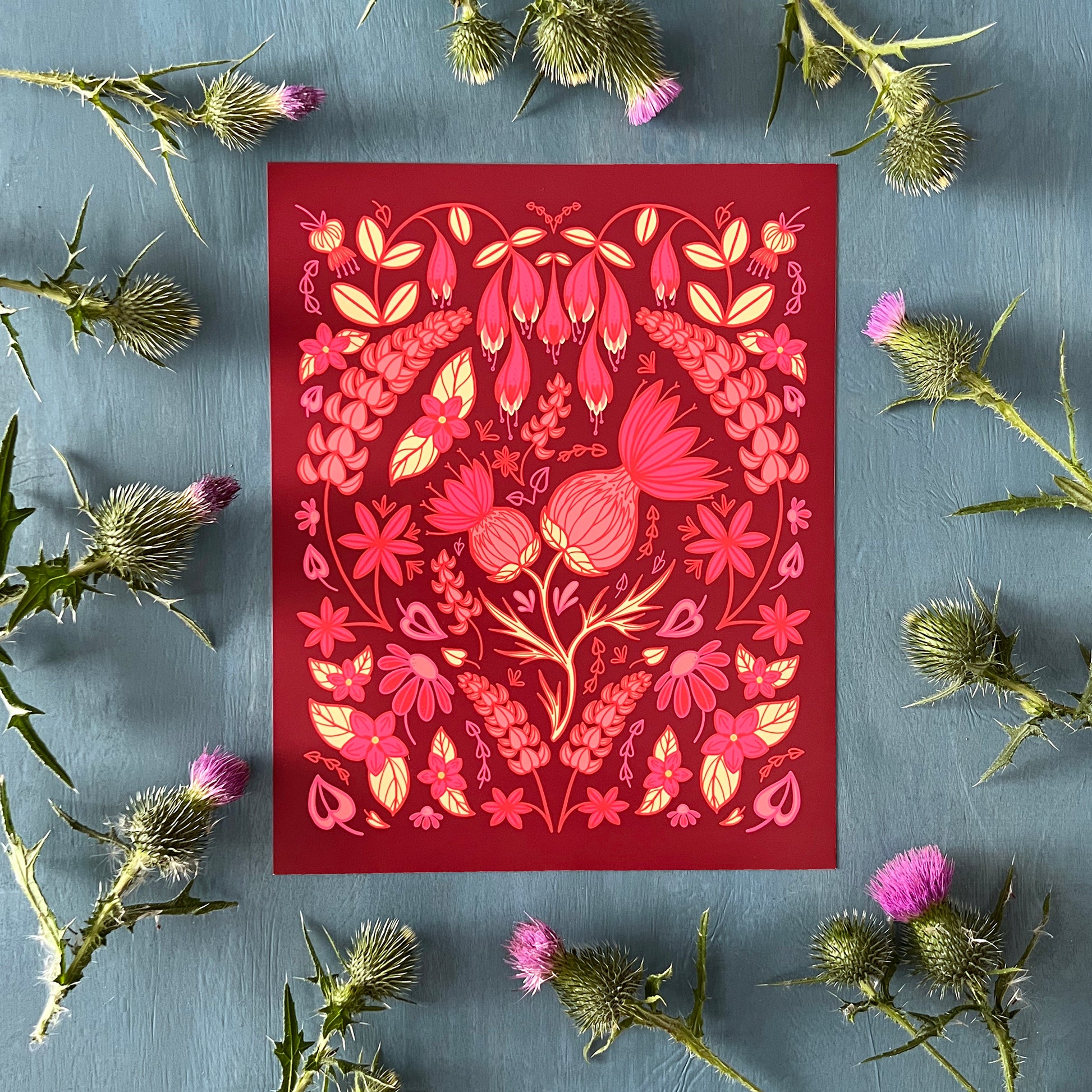 A pink and yellow illustration of thistles and other wildflowers in a full, folk-art style composition. Other flowers include lupies, echinacea, and haskap blossoms. The print is surrounded by thistles.