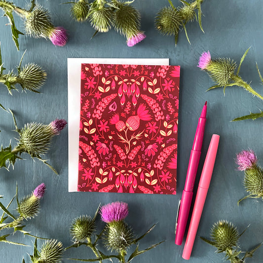 A dark pink greeting card is covered in a folk-style pattern of pink thistles, lupines, haskap berries and mayflowers, with creamy yellow leafy accents. The card is flanked by thistles and pink pens.