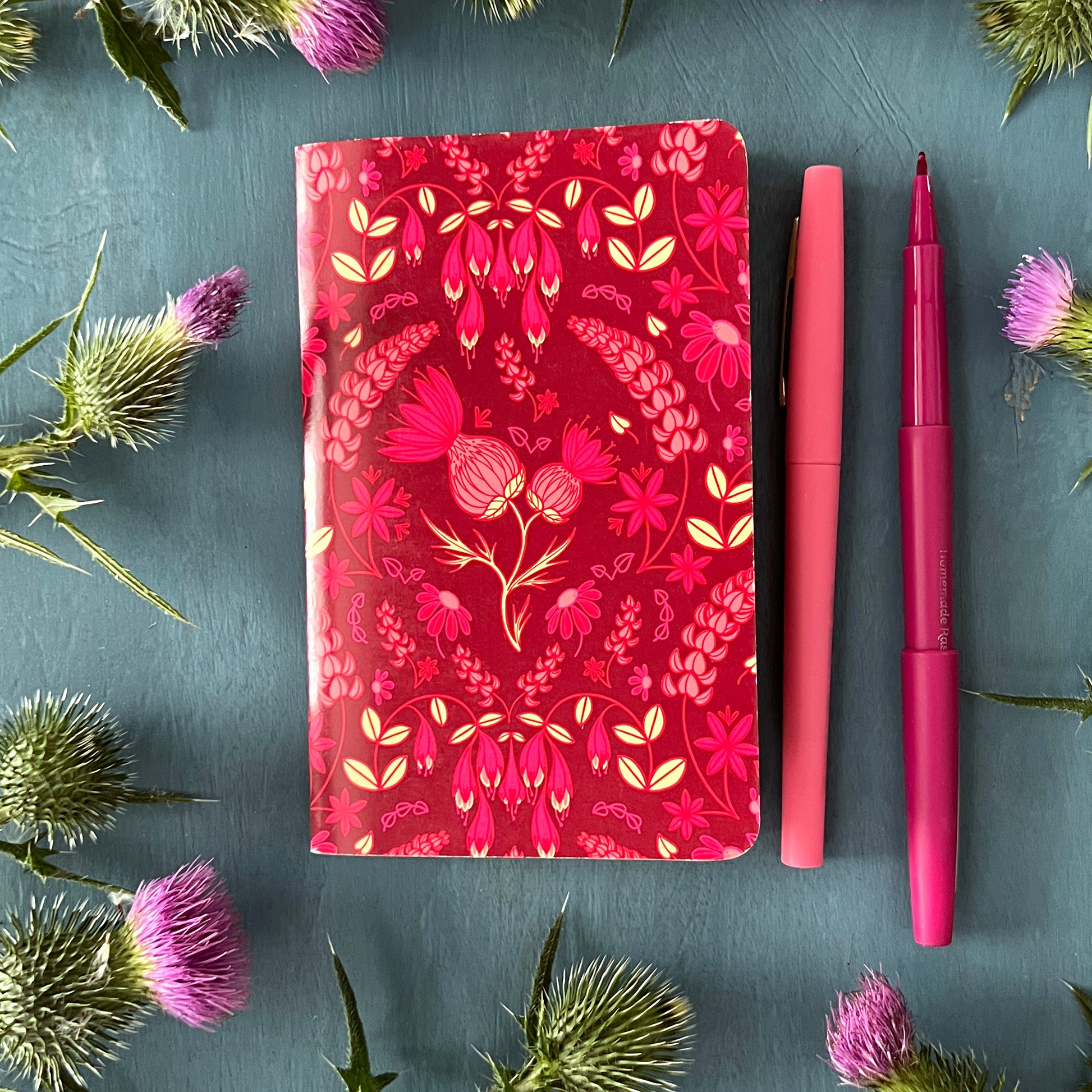 A pocket notebook with a thistle floral pattern on its cover rests next to pens and thistles.