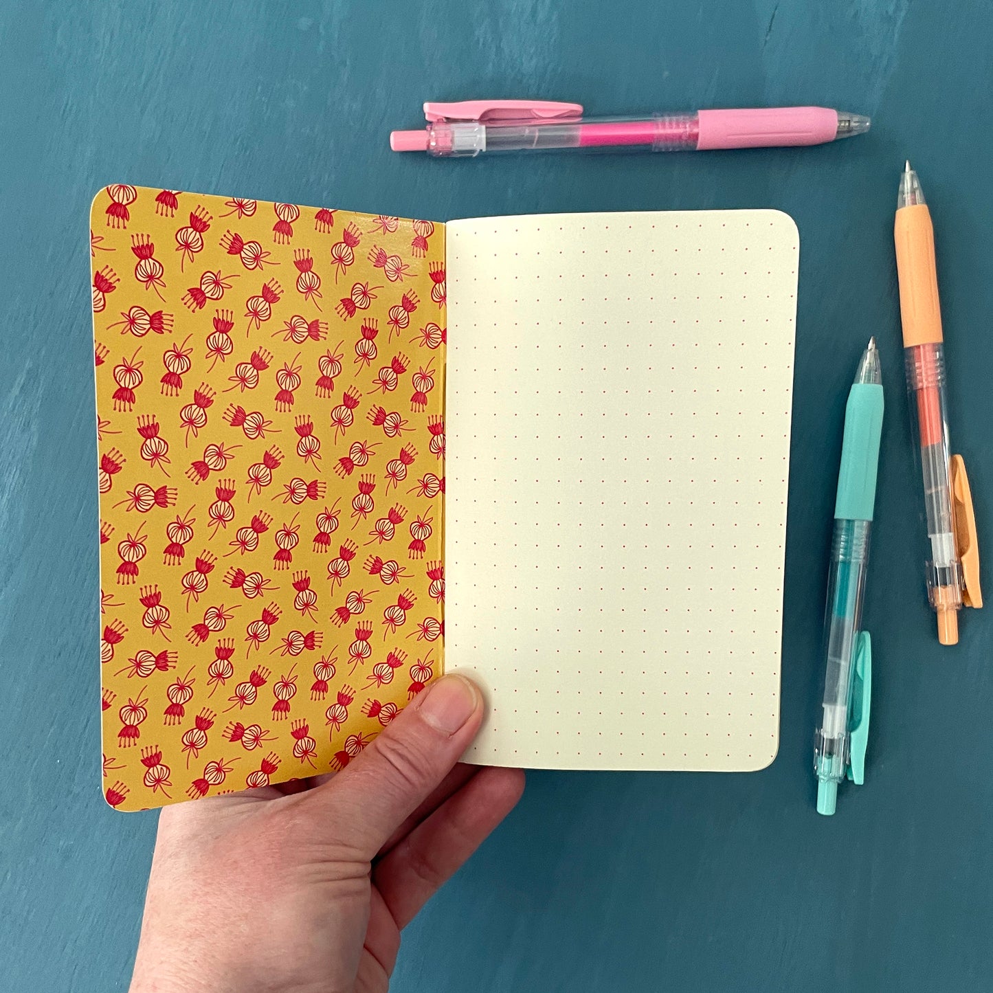 A pocket-sized notebook is open to its first page. The inner cover is yellow and patterned with stylized thistle blossoms. The notebook's first page is a dot grid in deep burgundy against creamy paper stock. The notebook is on a blue background surrounded by pens.