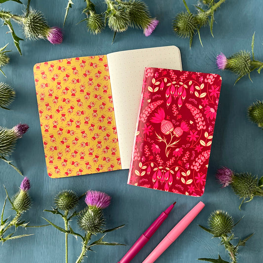 A pocket notebook with a thistle floral pattern on its cover rests on a notebook open to a dot grid page with a thistle scatter pattern on the inside cover. The notebooks are surrounded by thistles.
