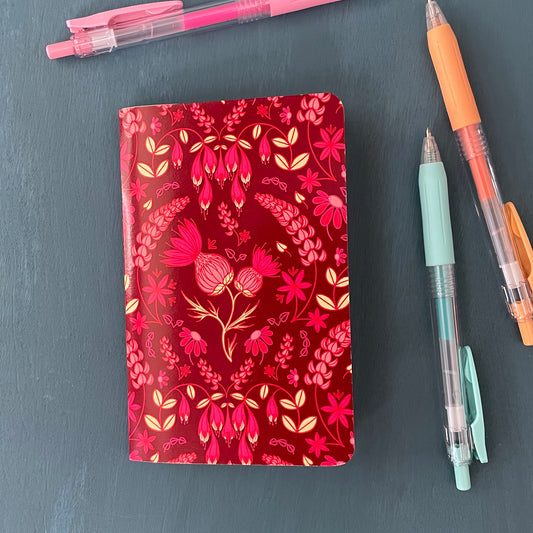 A pocket-sized notebook has a bright burgundy cover patterned with a folk floral made up of thistles, lupines, haskap blossoms and echinacea in shades of pink. It sits on a blue background surrounded by pens.