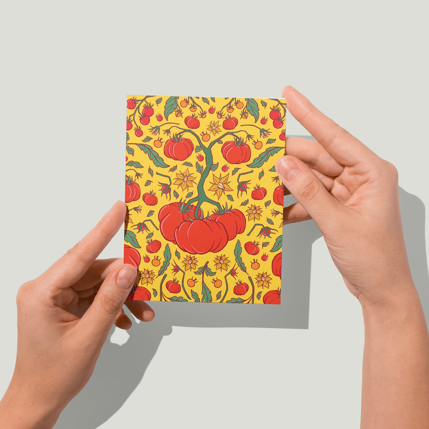 A card shows a yellow, green and red pattern of heirloom tomatoes, tomato flowers, and tomato leaves. A pair of hands holds the card up to the camera.