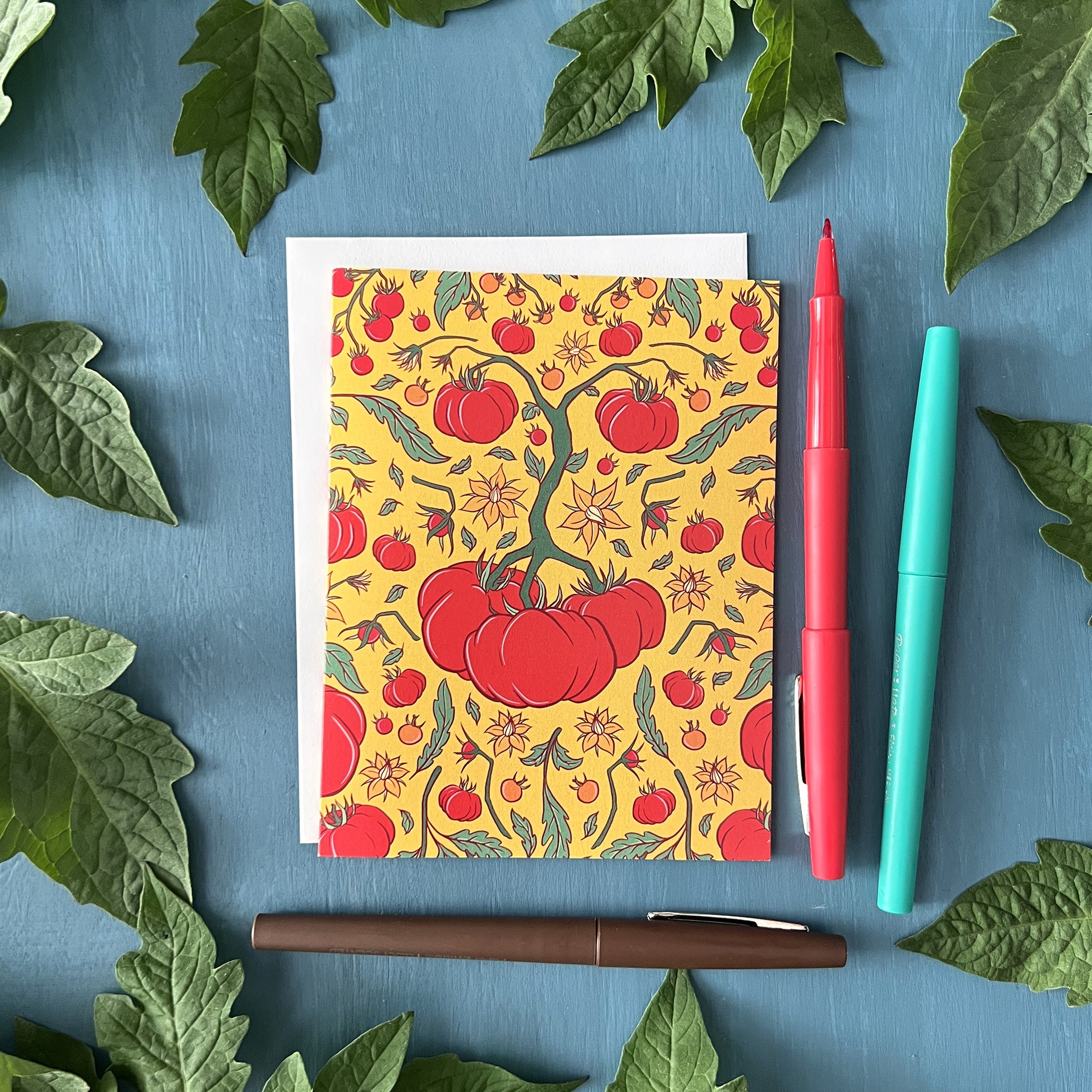 A card shows a yellow, green and red pattern of heirloom tomatoes, tomato flowers, and tomato leaves. The card is against a white envelope on a blue background surrounded by tomato leaves and pens.