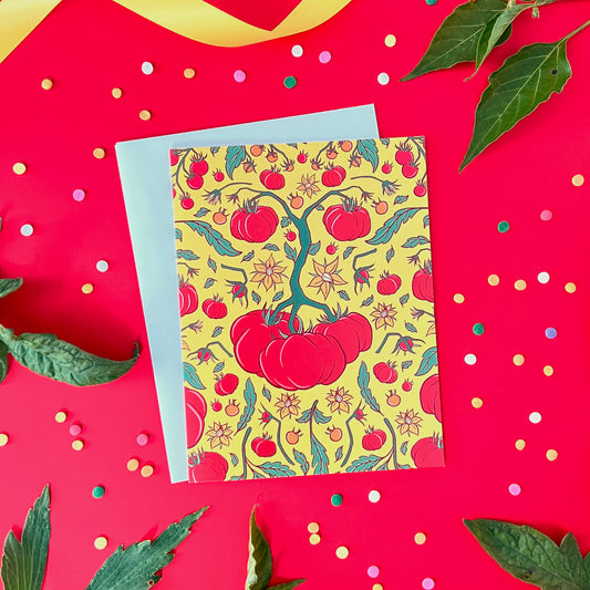 A yellow greeting card with a tomato pattern on it is on red surrounded by leaves and confetti.