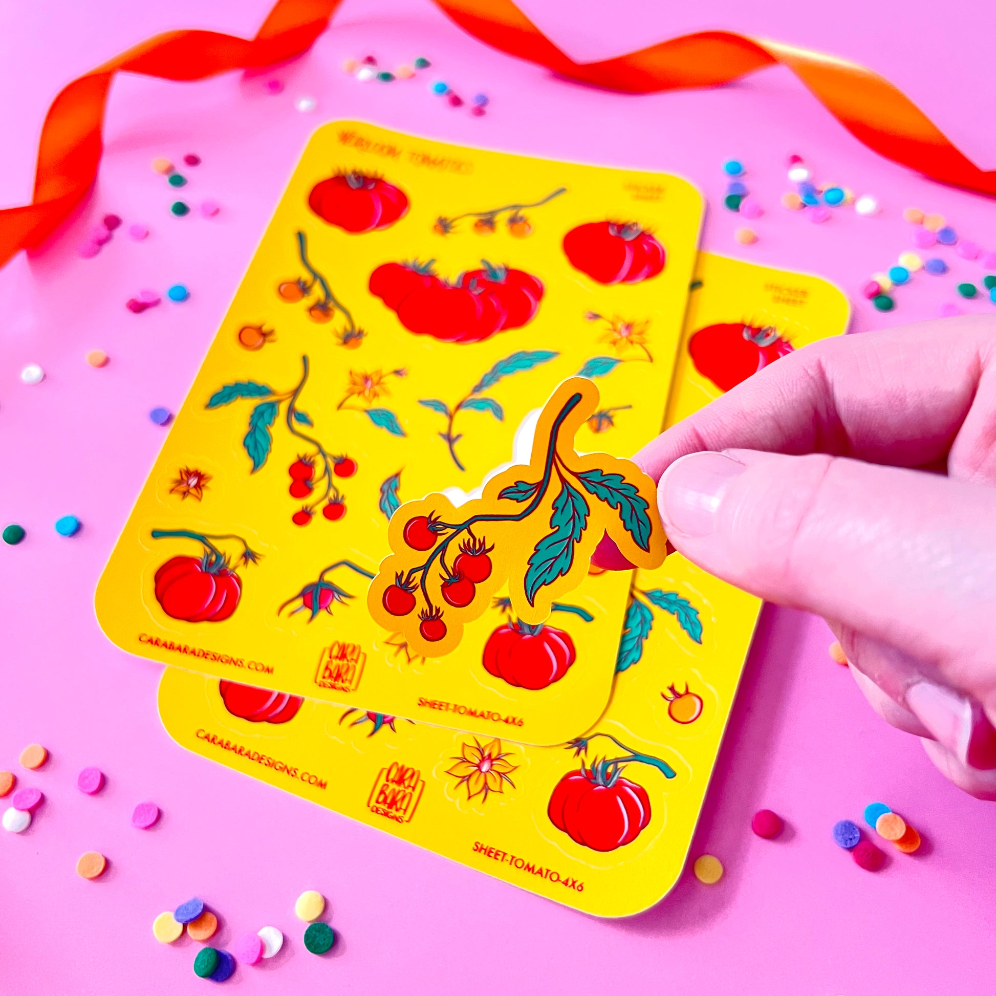 Two yellow sheets of tomato themed stickers lie flat on a pink background and a hand holds a cherry tomato sticker.