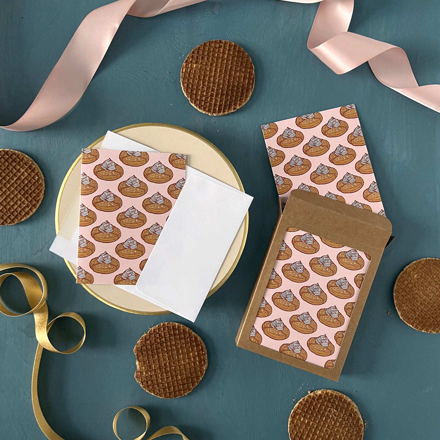 Pink greeting cards patterned with Belgian waffles appear on a blue background with pink ribbon and stroopwafels scattered around them.
