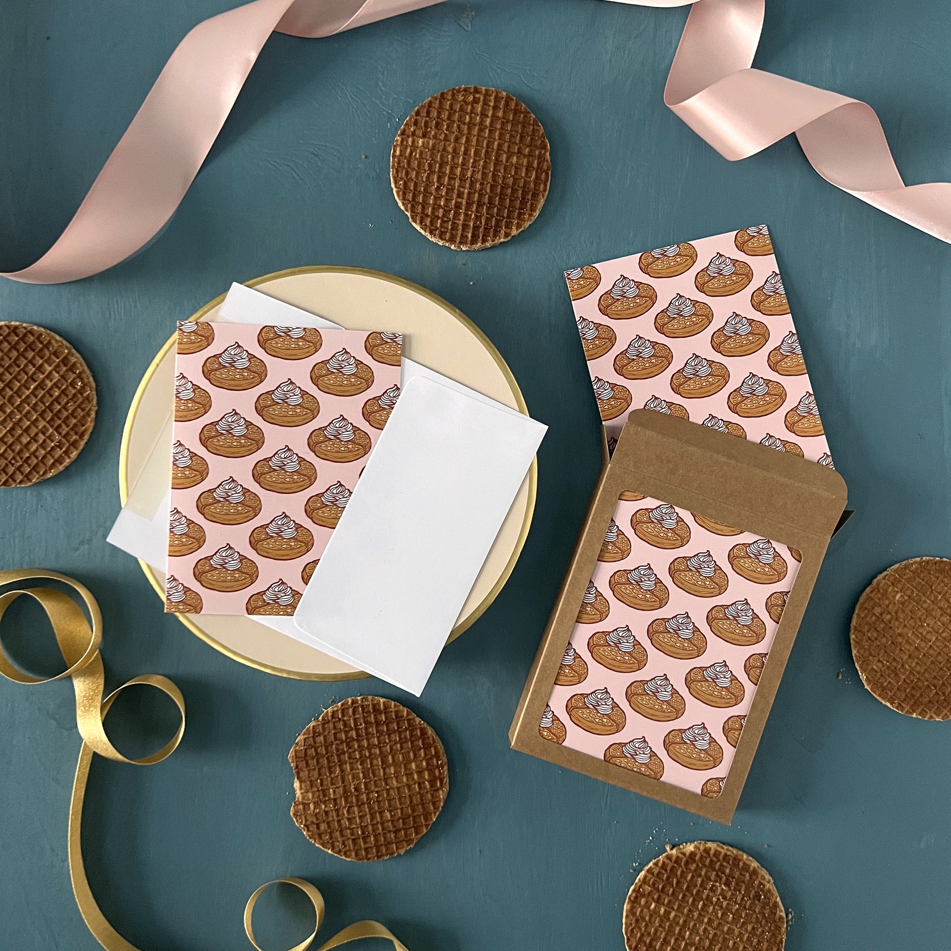 Pink greeting cards patterned with Belgian waffles appear on a blue background with pink ribbon and stroopwafels scattered around them.