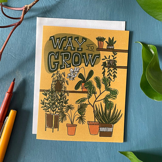 A mustard-yellow greeting card features illustrations of several potted plants, including a fiddle-leaf fig, a fan palm, ivy, and aloe plant, and an orchid. Green midcentury-inspired hand-lettering spells the words Way to Grow at the top of the card. The card sits against a blue wooden background and is flanked by leaves, vines and felt-tip pens.