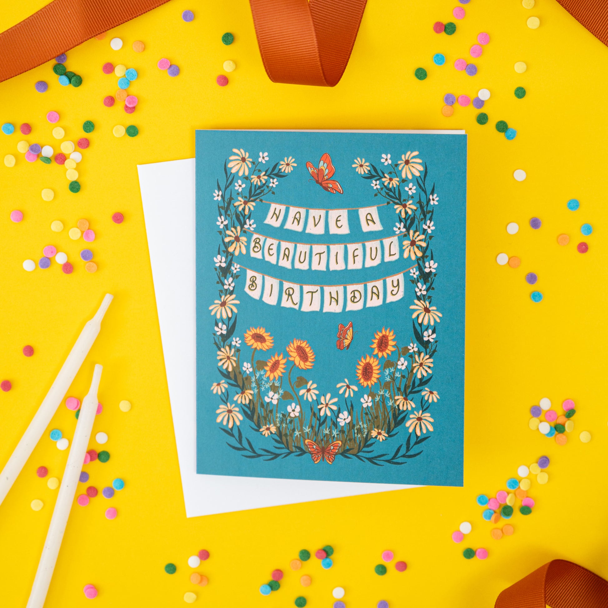 A blue birthday card shows an overgrown wildflower garden, butterflies, and the words 'have a beautiful birthday.' The card is on a yellow background surrounded by confetti and ribbon.