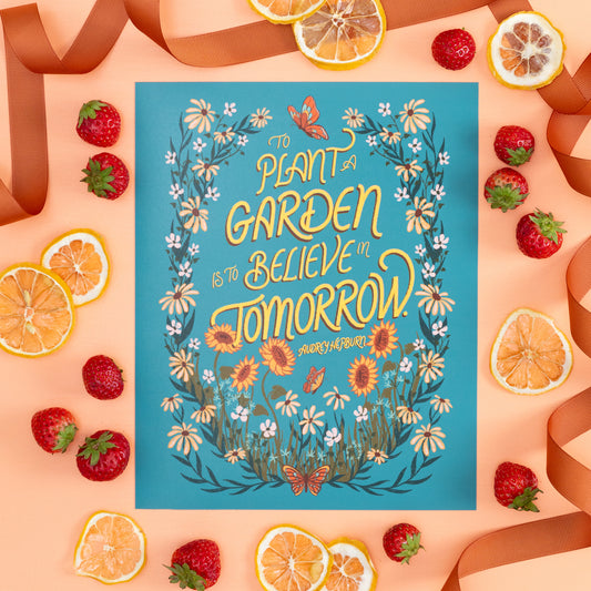 A hand lettered floral art print reads 'To plant a garden is to believe in tomorrow'. The print is on orange with strawberries and lemon slices.