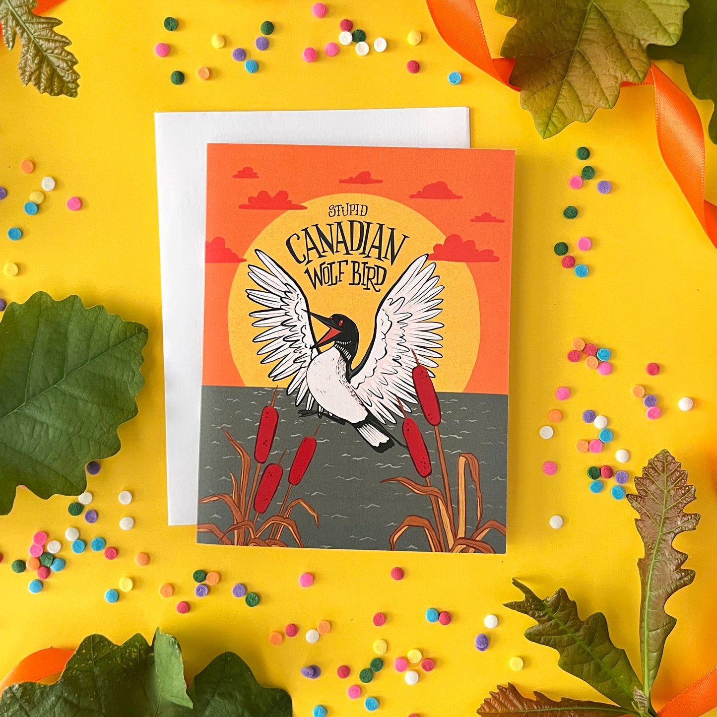 A greeting card with a loon says 'stupid Canadian Wolf Bird' in hand lettering. The card is with oak leaves and confetti on yellow.