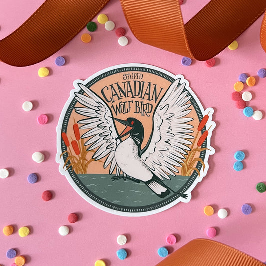 A vinyl sticker includes the words Stupid Canadian Wolf Bird and a squawking loon emerging from cattails on a lake. It is on pink with ribbon and confetti.