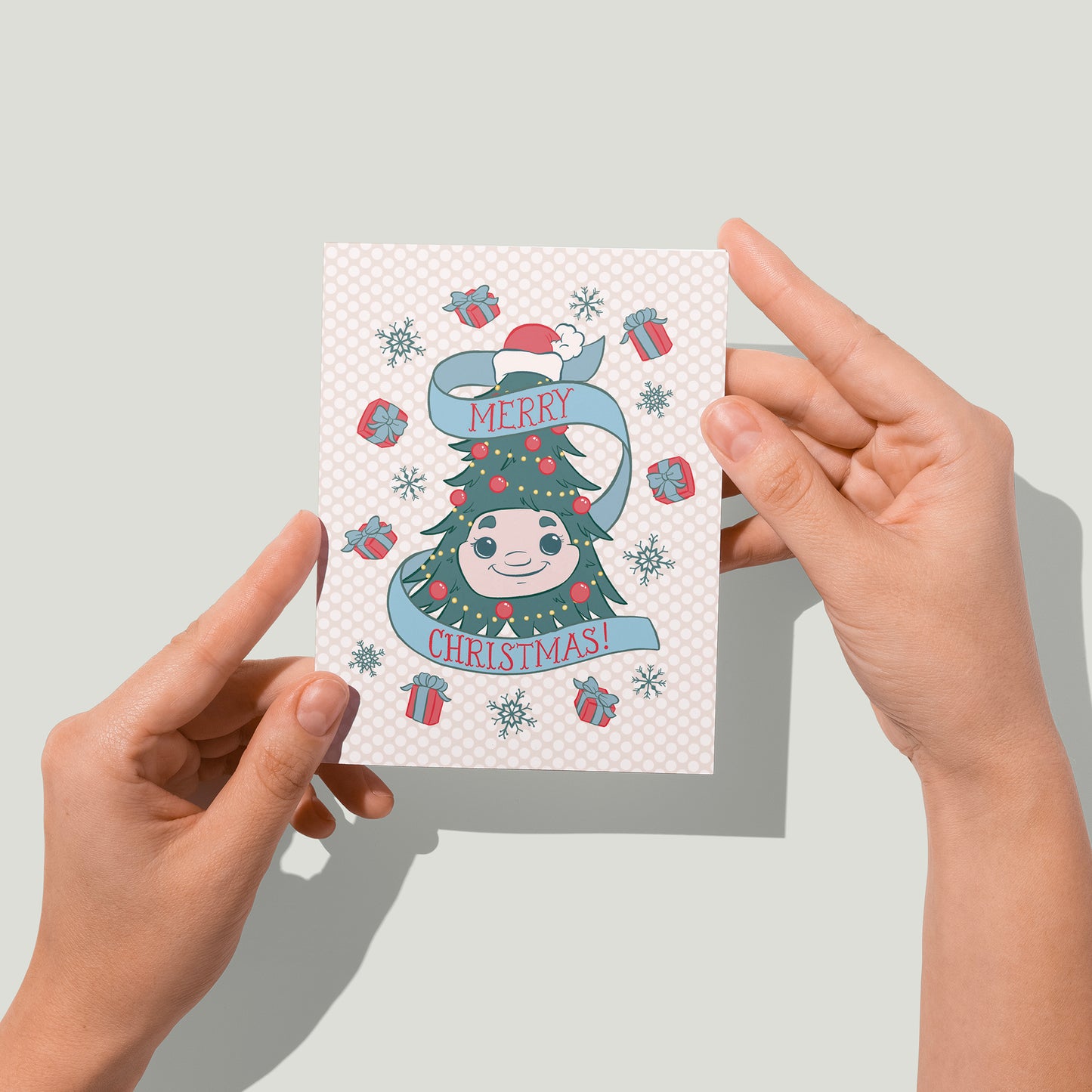 A Christmas card features an illustration of Woody the Christmas Tree, some gifts with bows and blue snowflakes on a pink dotted background. A banner reads Merry Christmas in hand-lettering. A pair of hands holds the card up in front of the camera.