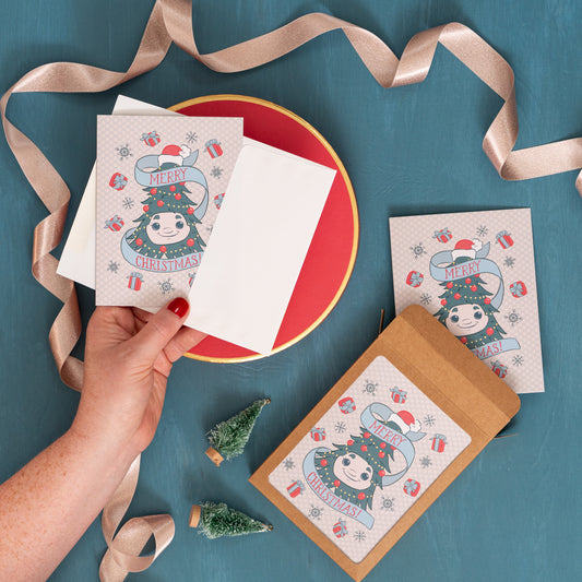 A hand holds a holiday greeting card with Woody the Christmas tree on it against a festive blue background with pink ribbon, bottle brush trees, and a box of the same card.