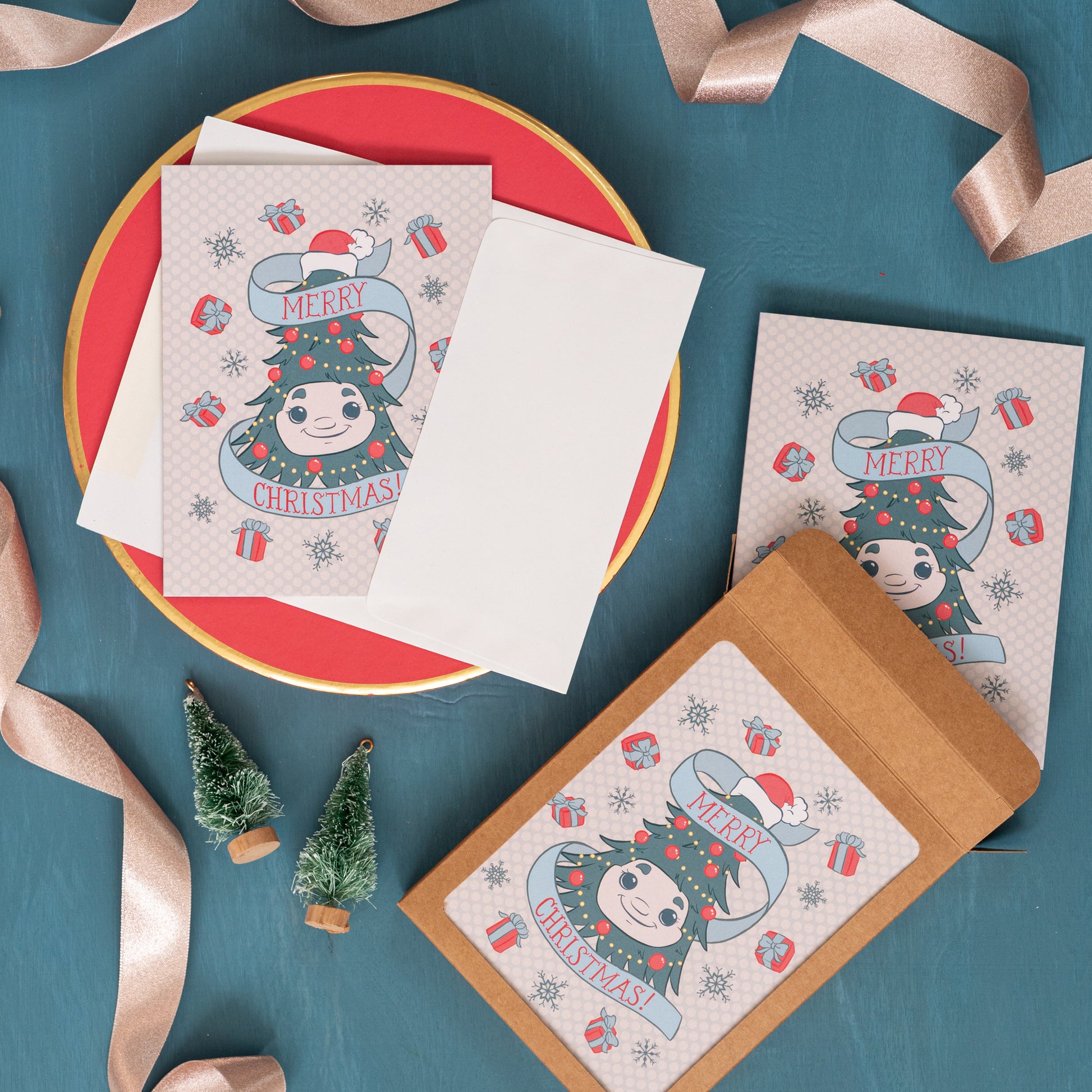 A holiday greeting card with Woody the Christmas tree on it appears against a festive blue background with pink ribbon, bottle brush trees, and a box of the same card.