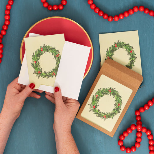 Hands hold a pale yellow Christmas card with an evergreen wreath on a festive blue background with red beads and a box of the same holiday card.