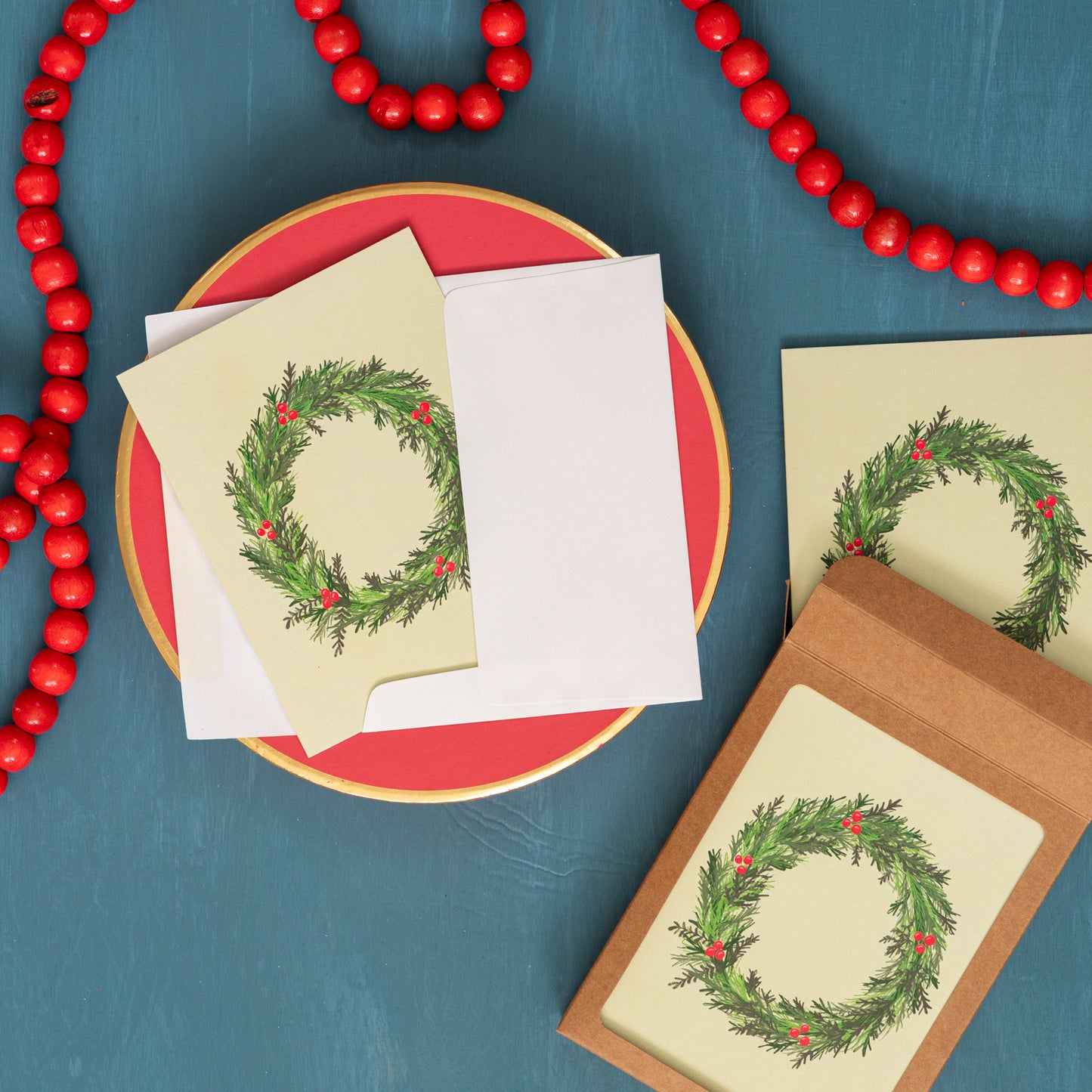 A pale yellow Christmas card with an evergreen wreath appears on a festive blue background with red beads and a box of the same holiday card.