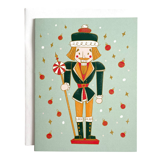 A mint card features a midcentury-style nutcracker in green and yellow, flanked by dots, yellow diamonds and red baubles. The card is against a white envelope on a white background.