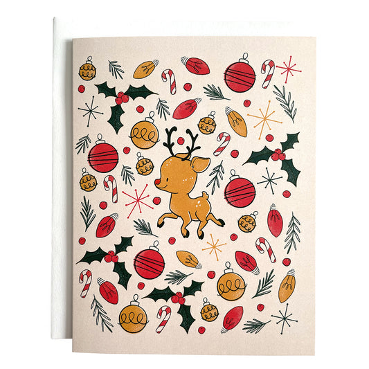 A blush pink card features a cute reindeer surrounded by an explosion of Christmas lights, ornaments, pine branches and holly. The card is against a white envelope on a white background.