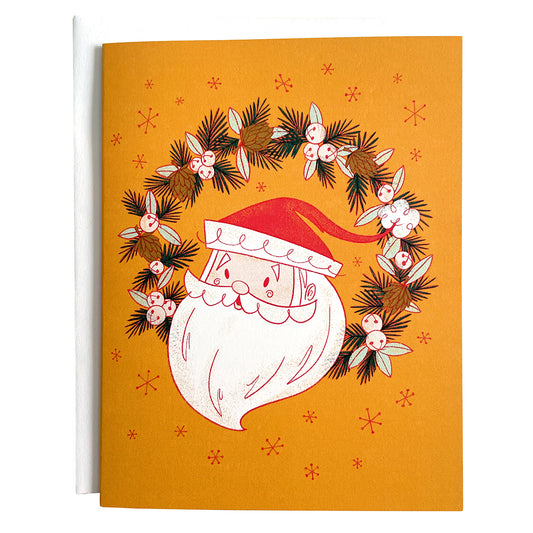 A yellow card shows a midcentury-style illustration of Santa's head in a wreath of jingle bells, pinecones and evergreen branches. The card is against a white envelope on a white background.