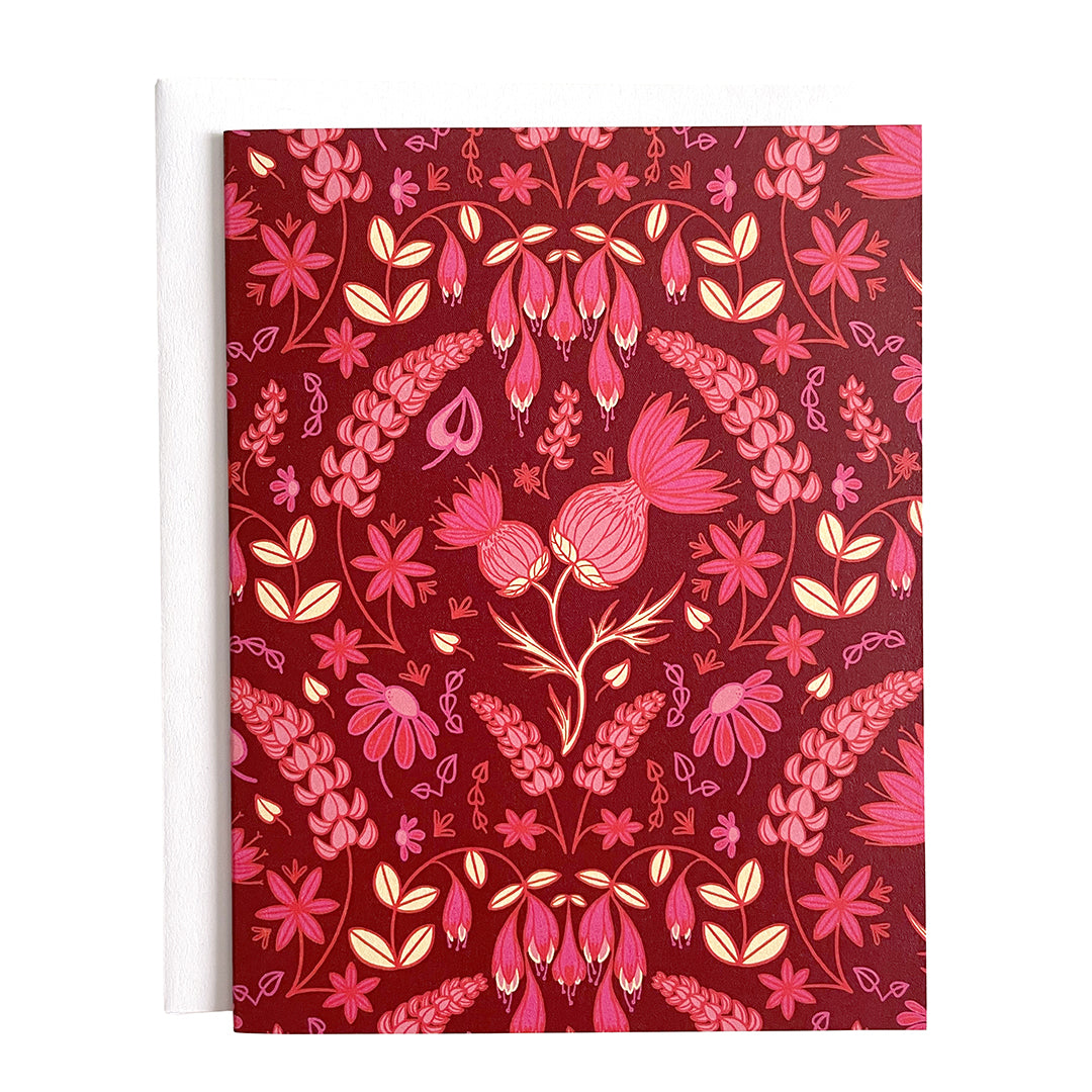 A dark pink greeting card is covered in a folk-style pattern of pink thistles, lupines, haskap berries and mayflowers, with creamy yellow leafy accents. The card is on a white envelope on a white background.