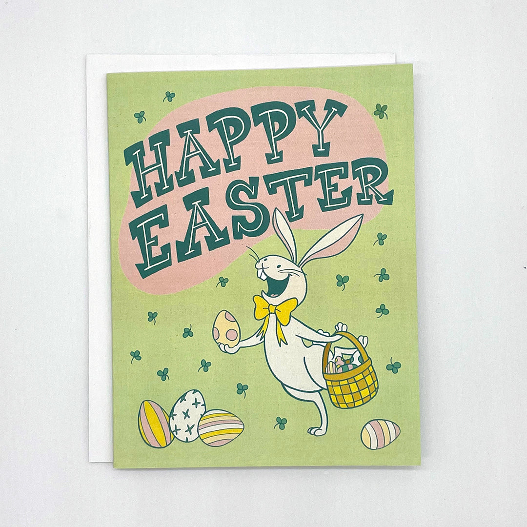 A pale green greeting card shows a cheerful white rabbit wearing a yellow bow tie holding a polka dotted easter egg in one paw and a basket of eggs in the other. There are easter eggs in front of and behind the rabbit. The card reads Happy Easter in midcentury-inspired hand-lettering and shows a shower of green clover. The card is on a white envelope on a white background.