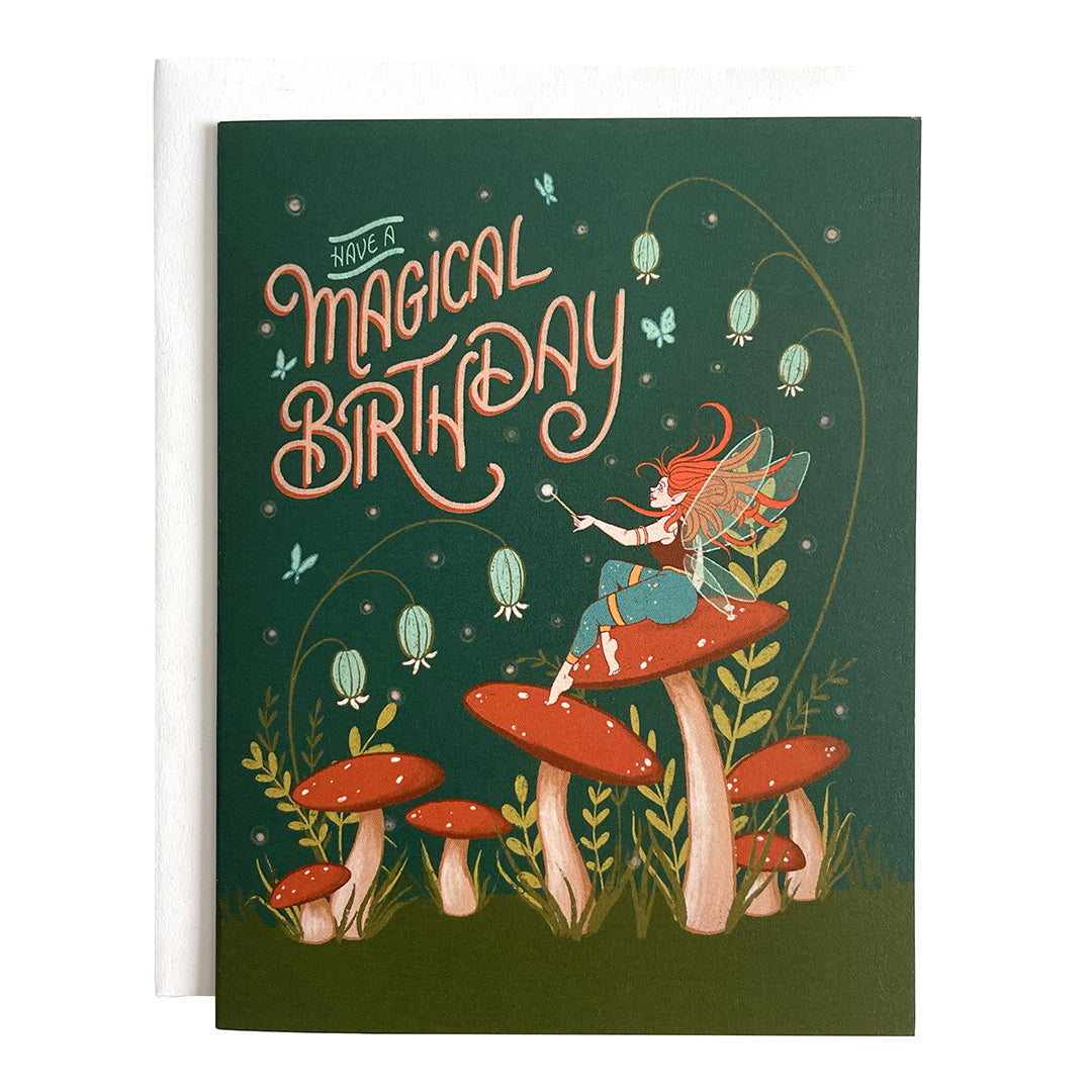 A dark green card says Have a Magical Birthday in blue letters. Below it on some grass are mushrooms, and a winged, red-haired faerie sits on one and looks up at the letters. Wildflowers appear behind the mushrooms. The card is on a white envelope on a white background.