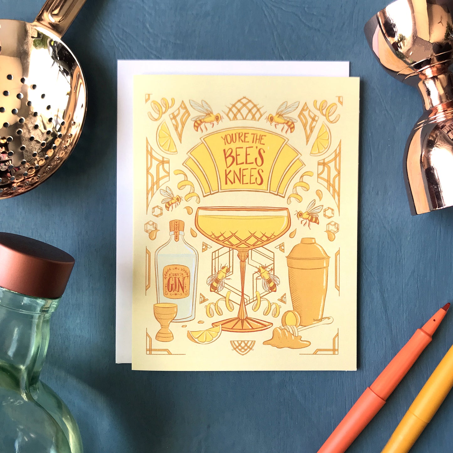 A yellow art-deco inspired greeting card is covered with illustrations of bees, honey and cocktail accessories, along with a crystal coupe filled with a Bee's Knees cocktail. A marquee on the card reads "You're the Bee's Knees" in dark orange letters. The card is surrounded by a bottle of Plymouth gin, a copper julep strainer and bell jigger, and felt tip pens on a blue wooden background.