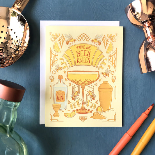A yellow art-deco inspired greeting card is covered with illustrations of bees, honey and cocktail accessories, along with a crystal coupe filled with a Bee's Knees cocktail. A marquee on the card reads You're the Bee's Knees in dark orange letters. The card is surrounded by a bottle of Plymouth gin, a copper julep strainer and bell jigger, and felt tip pens on a blue wooden background.