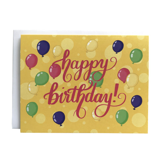 A yellow greeting card is covered with yellow dots and illustrations of pink, purple, yellow and green balloons. On the card are the words "happy birthday" lettered in bright pink. The card sits against a white envelope on a white background.